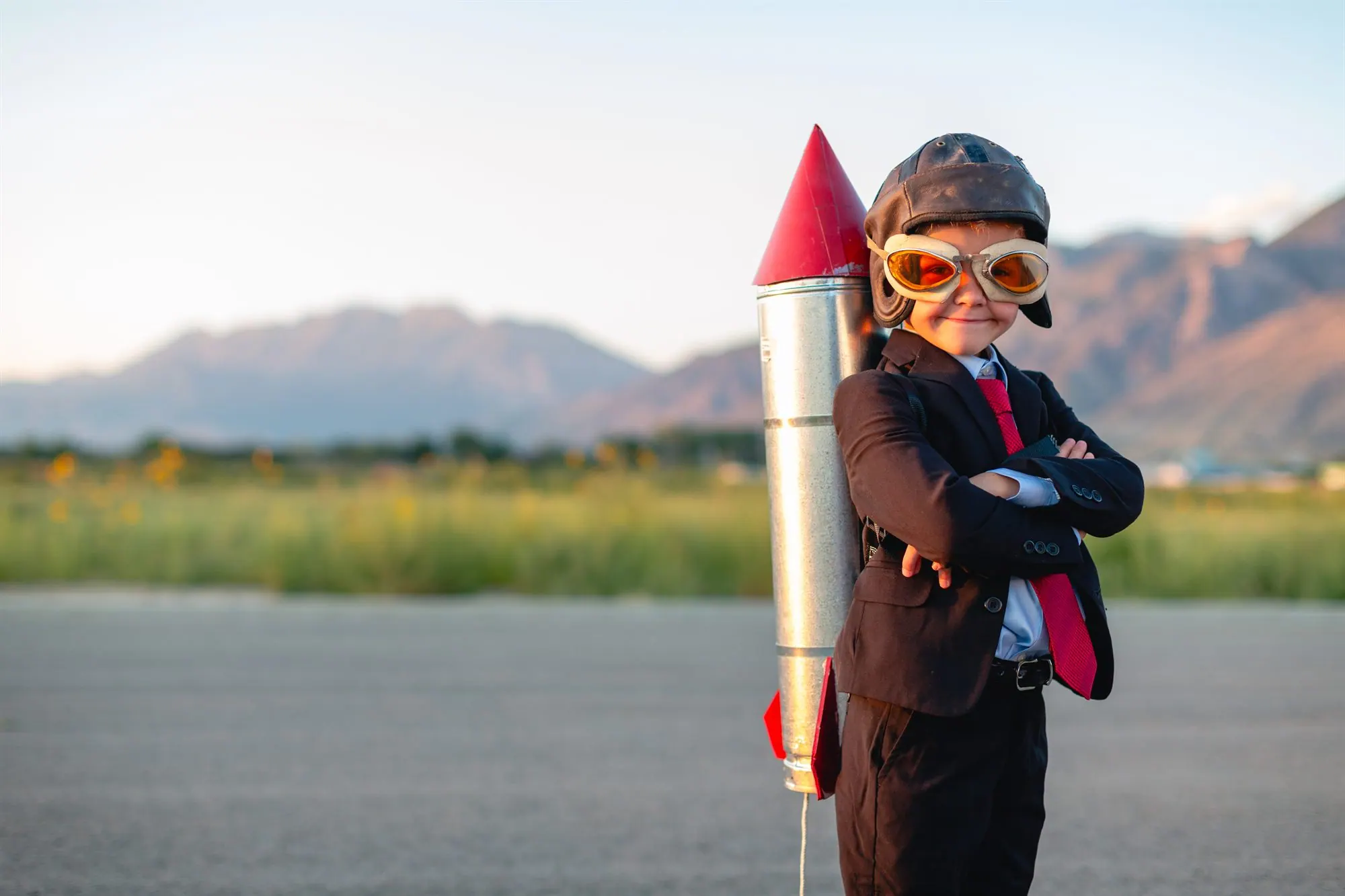 Kind im Anzug mit Fliegerbrille und selbstgebauter Rakete vor einer Bergkulisse — Sinnbild für große Ambitionen.